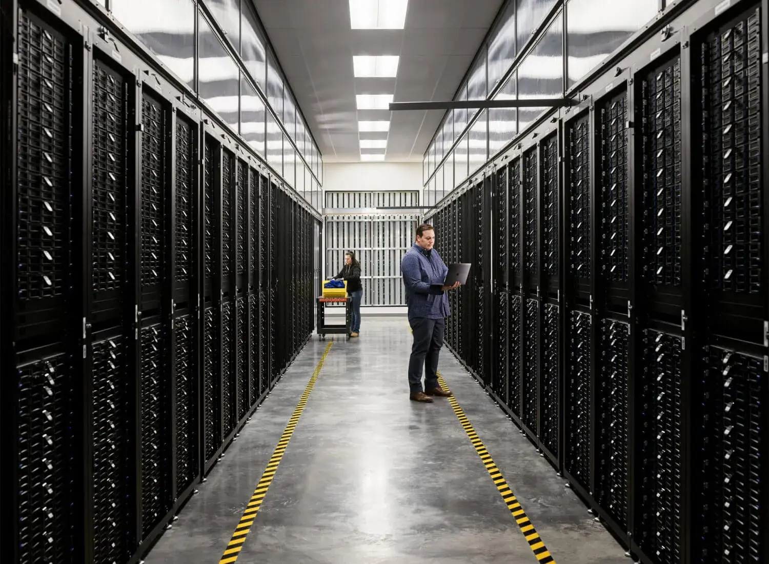 Apple IT Engineer with Laptop in between Server Aisles while a Technician Pushes Cart in the Background